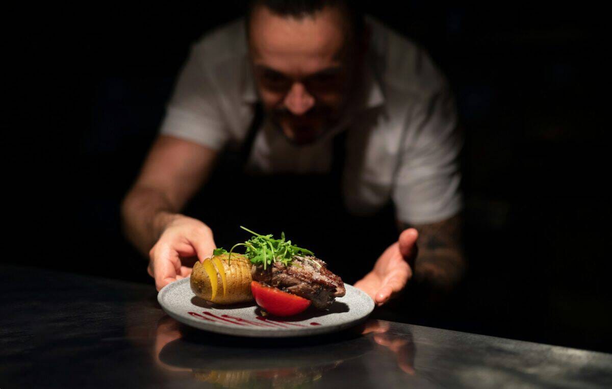 Chef handing plate with meal through order station in the commercial kitchen on black background