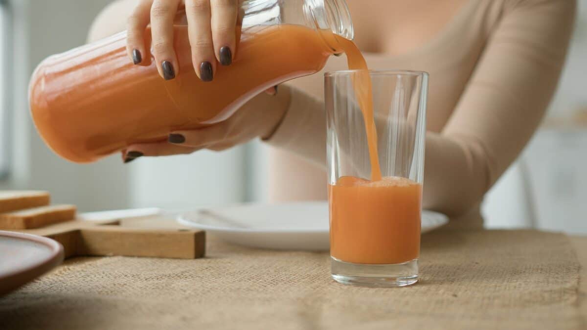 Close up unrecognizable woman pour orange juice from jar to glass on kitchen table. Female hands