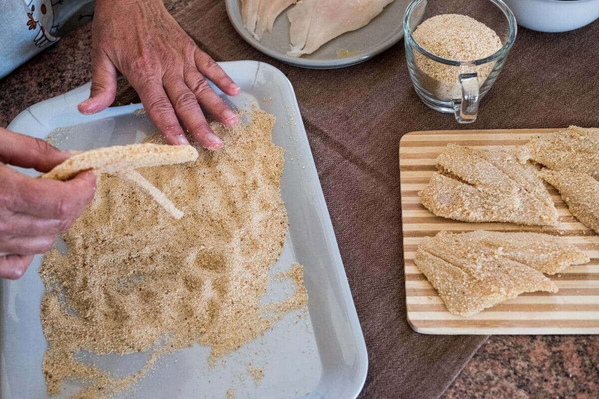 Elderly woman hands preparing breaded fish filets with eggs, wooden chopping board
