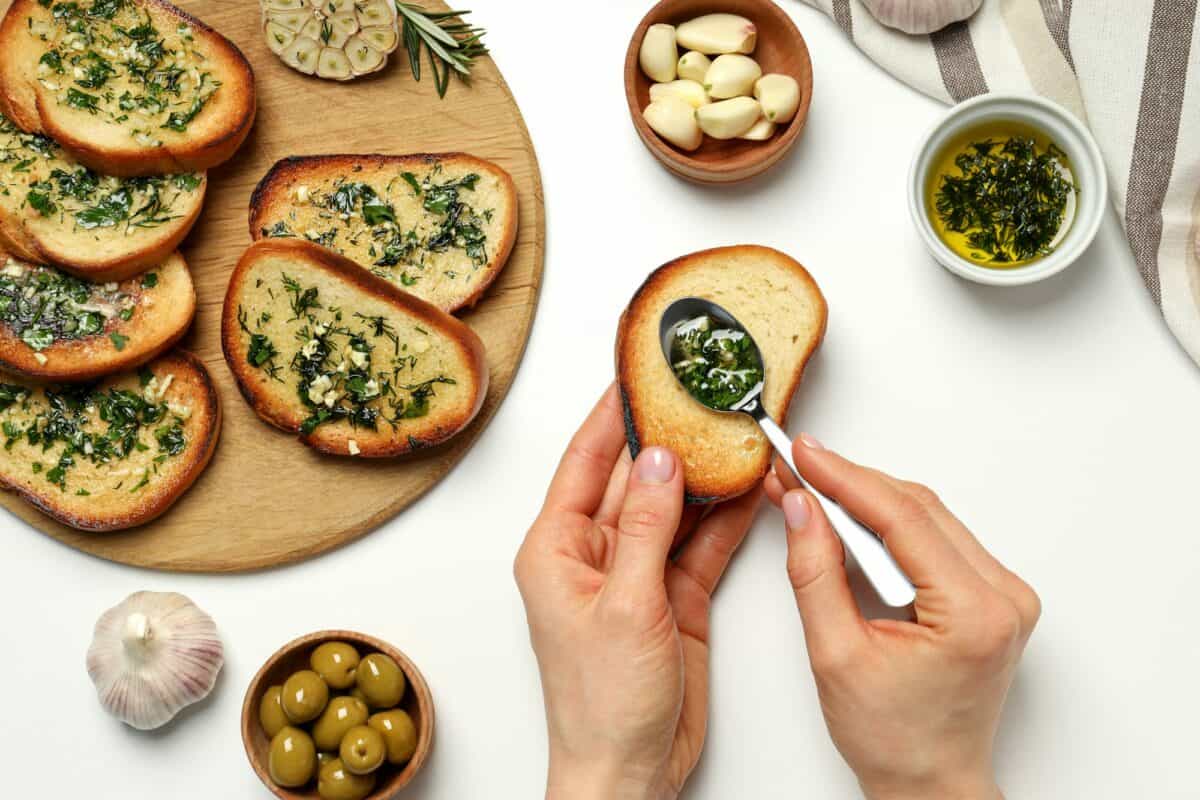 Garlic bread, hands, bowls with oil, olives and garlic on white background, top view