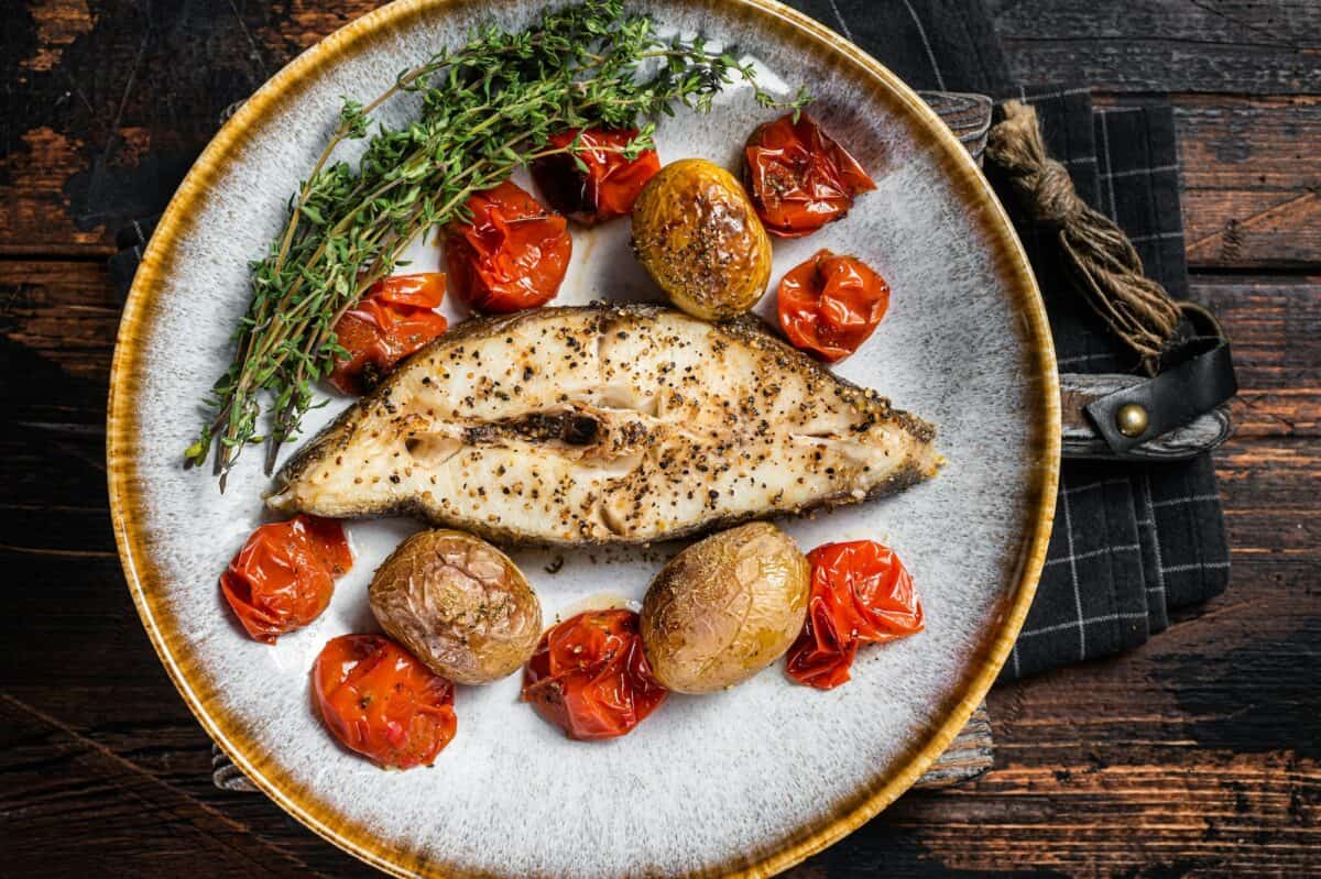 Grilled halibut fish steaks with tomato and potato in plate. Wooden background. Top view