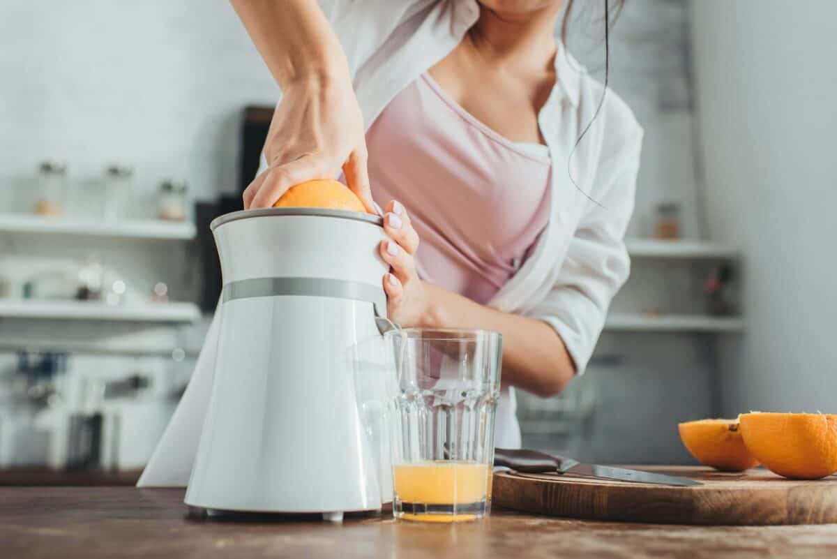 partial view of girl making fresh orange juice with juicer at wooden table in kitchen