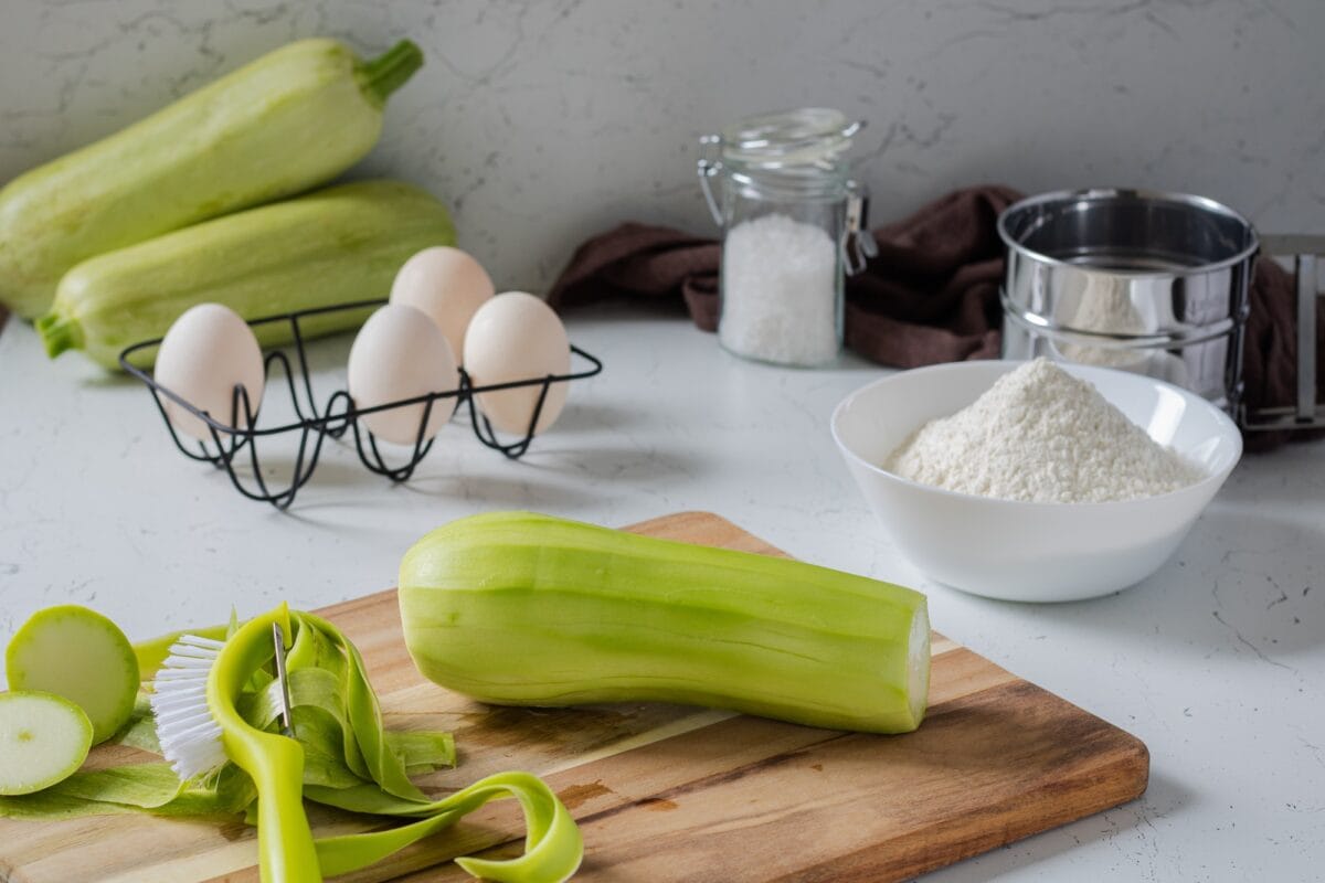 Peeled zucchini and a vegetable peeler on a board, next to flour, salt and eggs.