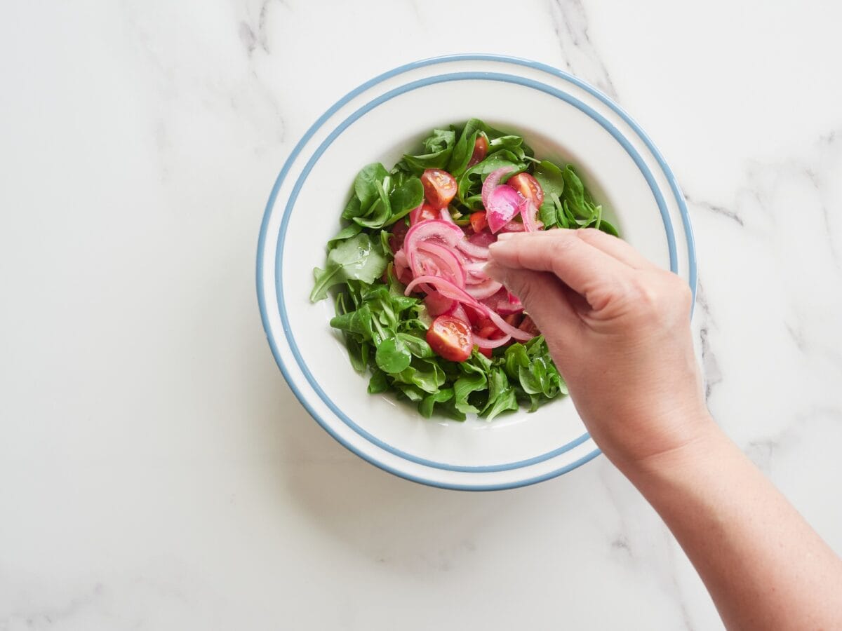 Top view of a person's hand dressing a plate of colorful salad with a pinch of salt.