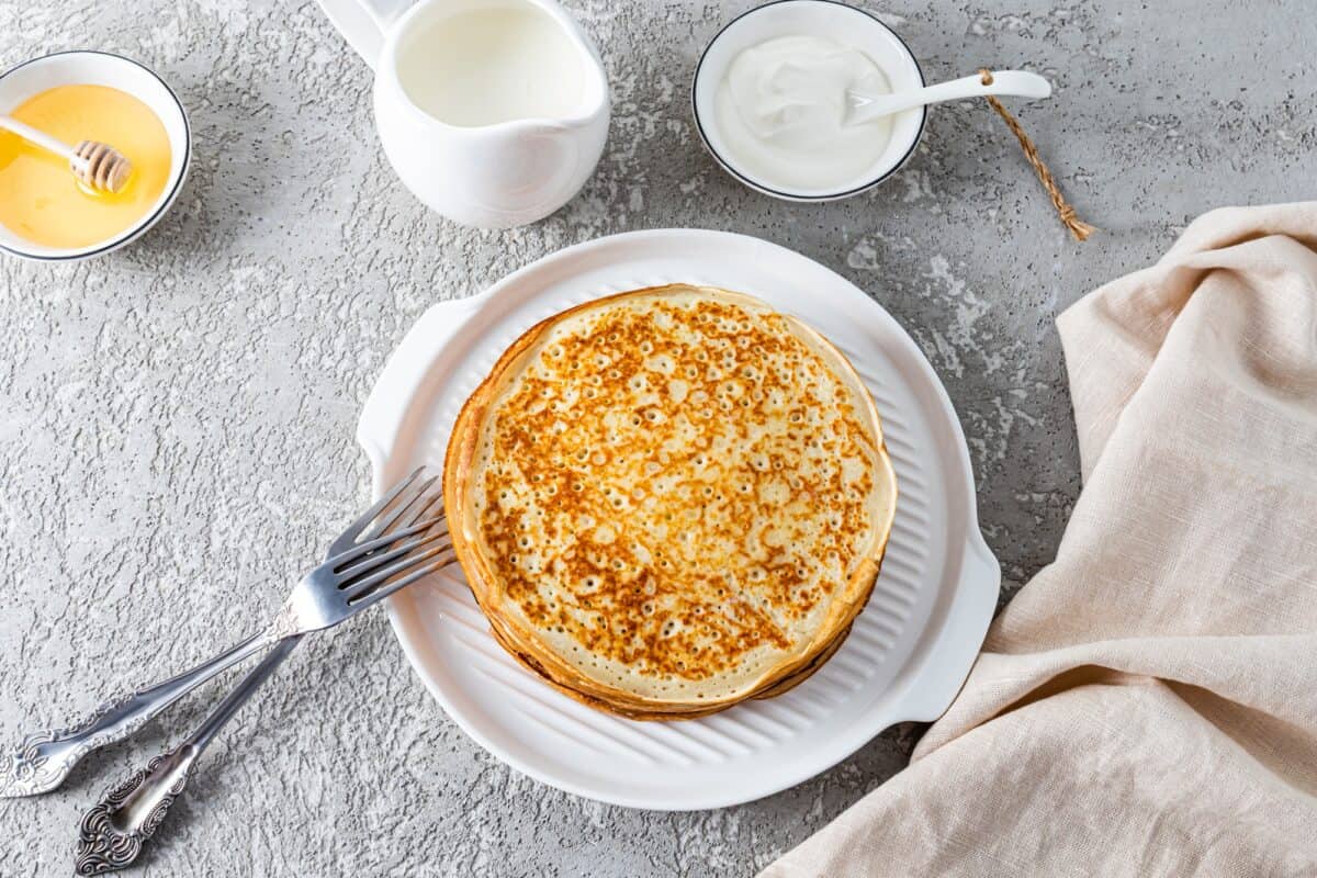 Vue de dessus d'une pile de délicieuses crêpes sur une assiette blanche, couverts, crème aigre, miel dans des bols